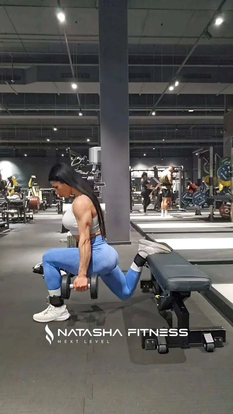 Natasha hace una zancada con el pie trasero apoyado en un banco y una mancuerna en la mano, en un gimnasio amplio