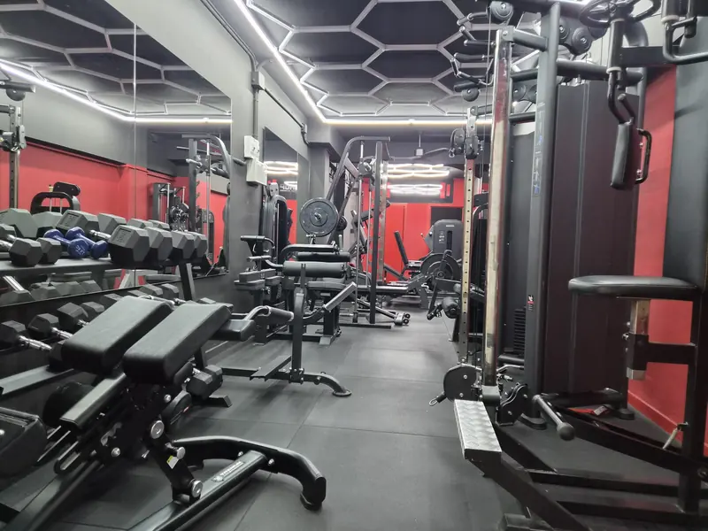 Sala de entrenamiento con suelo de caucho, rack de mancuernas, banco regulable, polea y paredes rojas, vista desde la entrada